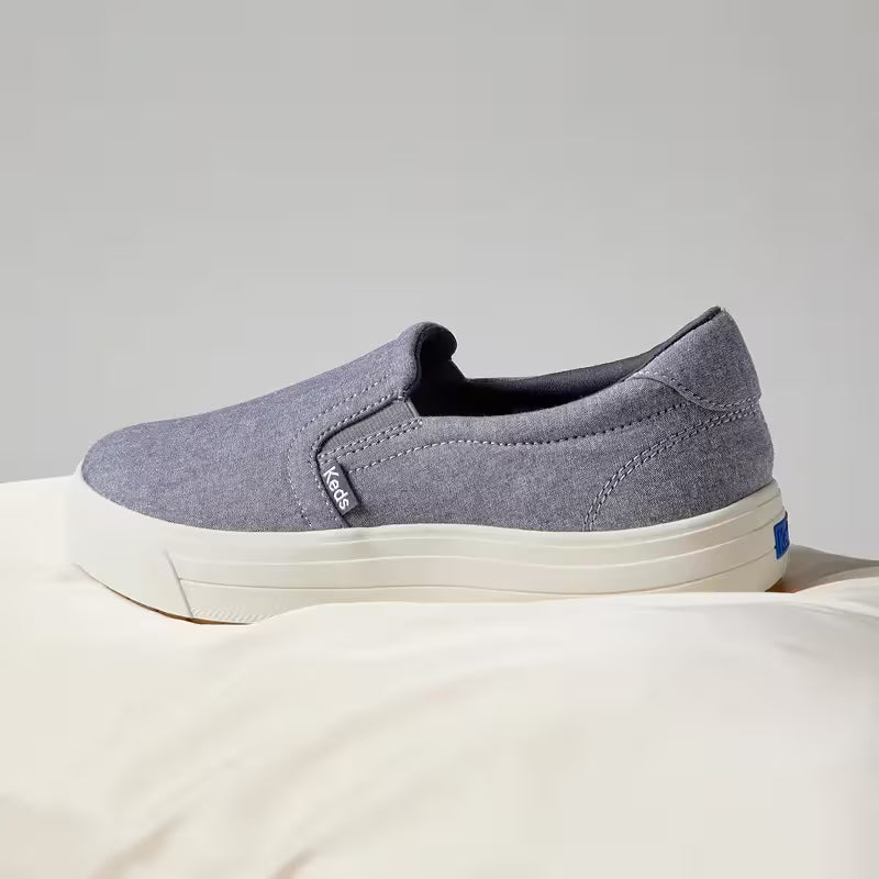A grey background with a side view of the Park Chambray Slip-On in blue.