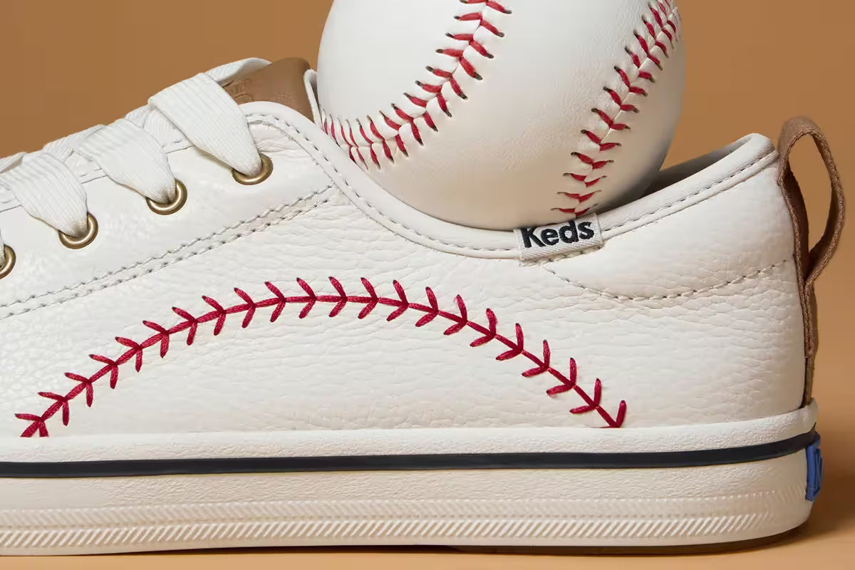 A tan background with a close-up side view of the Keds Champion Pennant Leather Lace-Up in off white with a baseball inside the shoe.