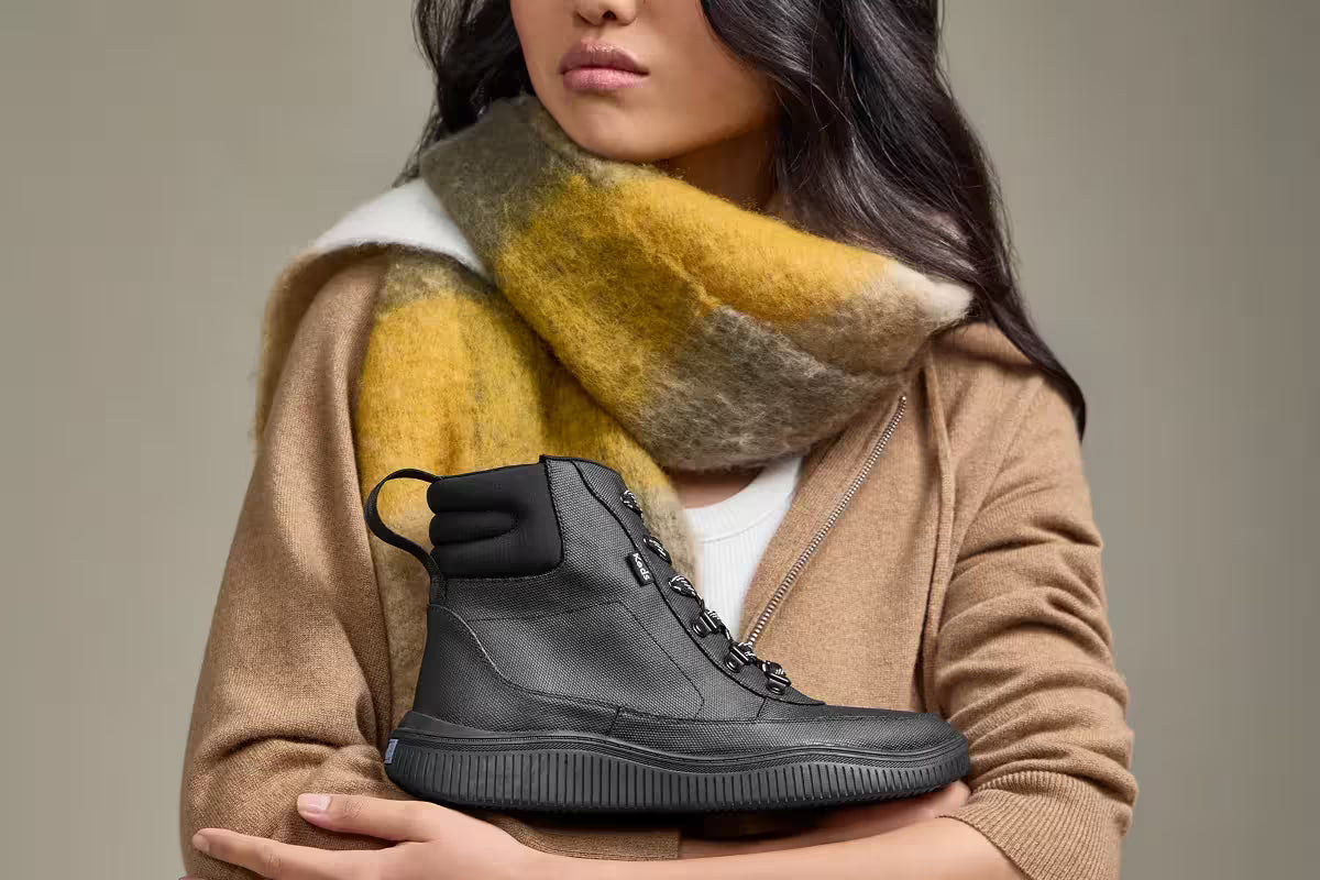 A grey background with a woman wearing a white T-shirt, chunky plaid scarf in autumn colors, and a tan zip-up sweatshirt. She is holding the Nova Coated Canvas/Nylon Lace-Up Boot.